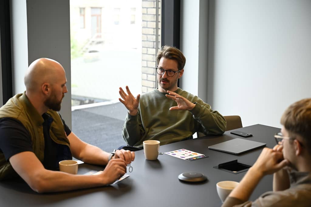 Man in green sweater speaking, gesturing to two colleagues at a modern meeting table.